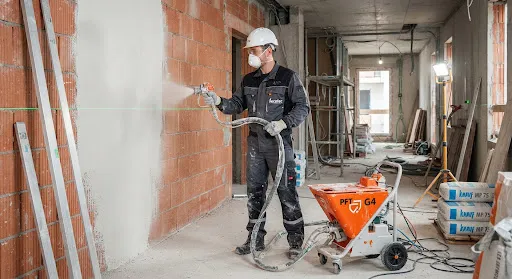 A construction worker wearing white safety helmet and dust mask operating a PFT G4 continuous plastering machine inside an apartment building, spraying smooth white gypsum plaster onto a red hollow brick wall guided by a sharp bright green laser level line, aluminum screed rulers standing against the wall, industrial construction site lighting, high realism architectural photography.